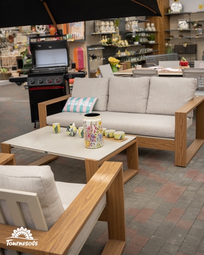 Ensemble de salon de jardin en bois clair et coussins beiges exposé dans un magasin avec un barbecue.